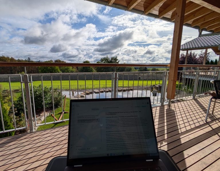 Schreiben Blick auf eine grüne Wiese und den Himmel von einer Terrasse mit Laptop im Vordergrund.